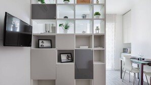 A modern living room with a flat-screen TV mounted on the wall, a dining area with a table and chairs, and a built-in shelving unit with decorative items and plants.