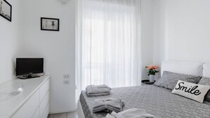 A modern bedroom with a bed, a TV on a dresser, and a window with sheer curtains.