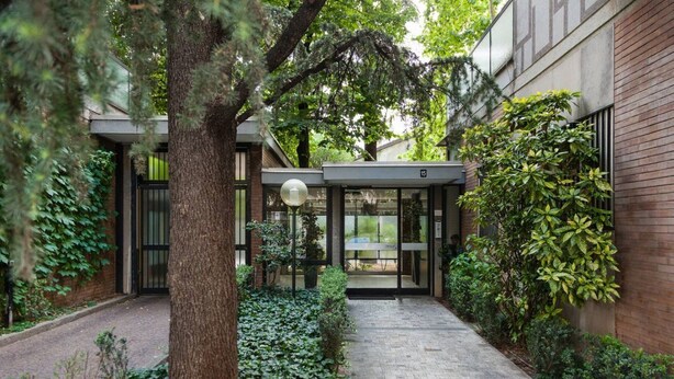 A modern building entrance with a glass door, surrounded by greenery and a paved path.
