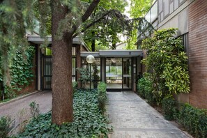 Entrada de un edificio moderno con puerta de cristal, rodeada de follaje y un sendero pavimentado.