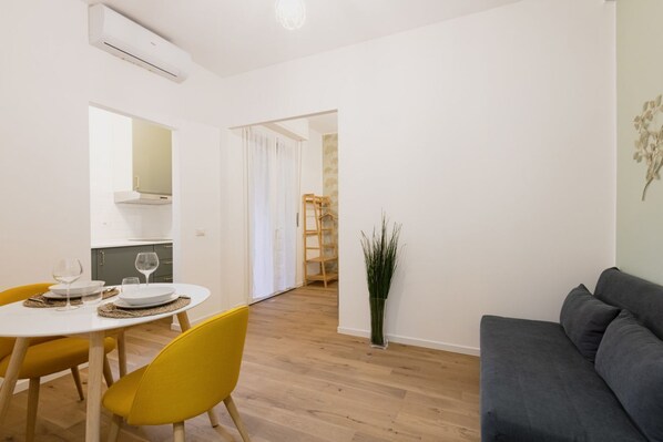 A modern living room with a gray sofa, a dining area with yellow chairs and a white table, and a kitchen visible through an open doorway. - Easylife - Stylish abode a few steps from Navigli (Milano)