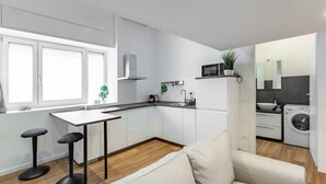 Une cuisine moderne avec des armoires blanches, un plan de travail noir et un four à micro-ondes encastré. On y trouve une fenêtre avec des stores blancs, un évier et un lave-linge dans un coin.