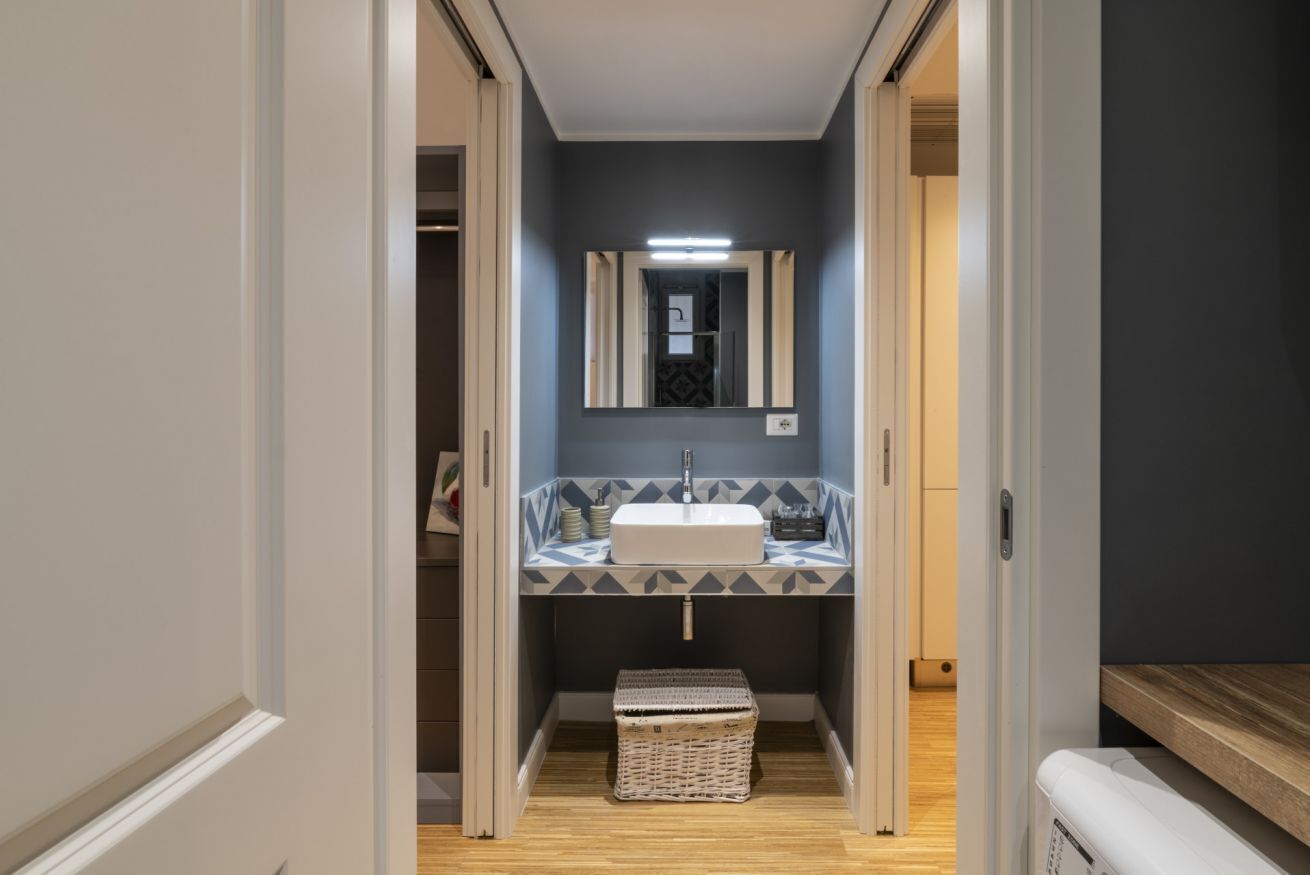 Un couloir étroit mène à une salle de bain équipée d’un lavabo blanc, d’un miroir et d’un pare-chocs décoré de motifs.