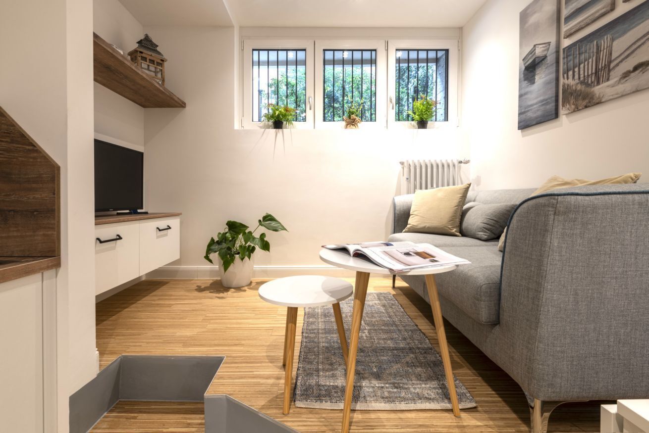 Ein kompakter Wohnraum mit grauem Sofa, einem kleinen runden Tisch mit einem Buch, einem Fernseher auf einem Holzregal und Topfpflanzen in der Nähe der Fenster.