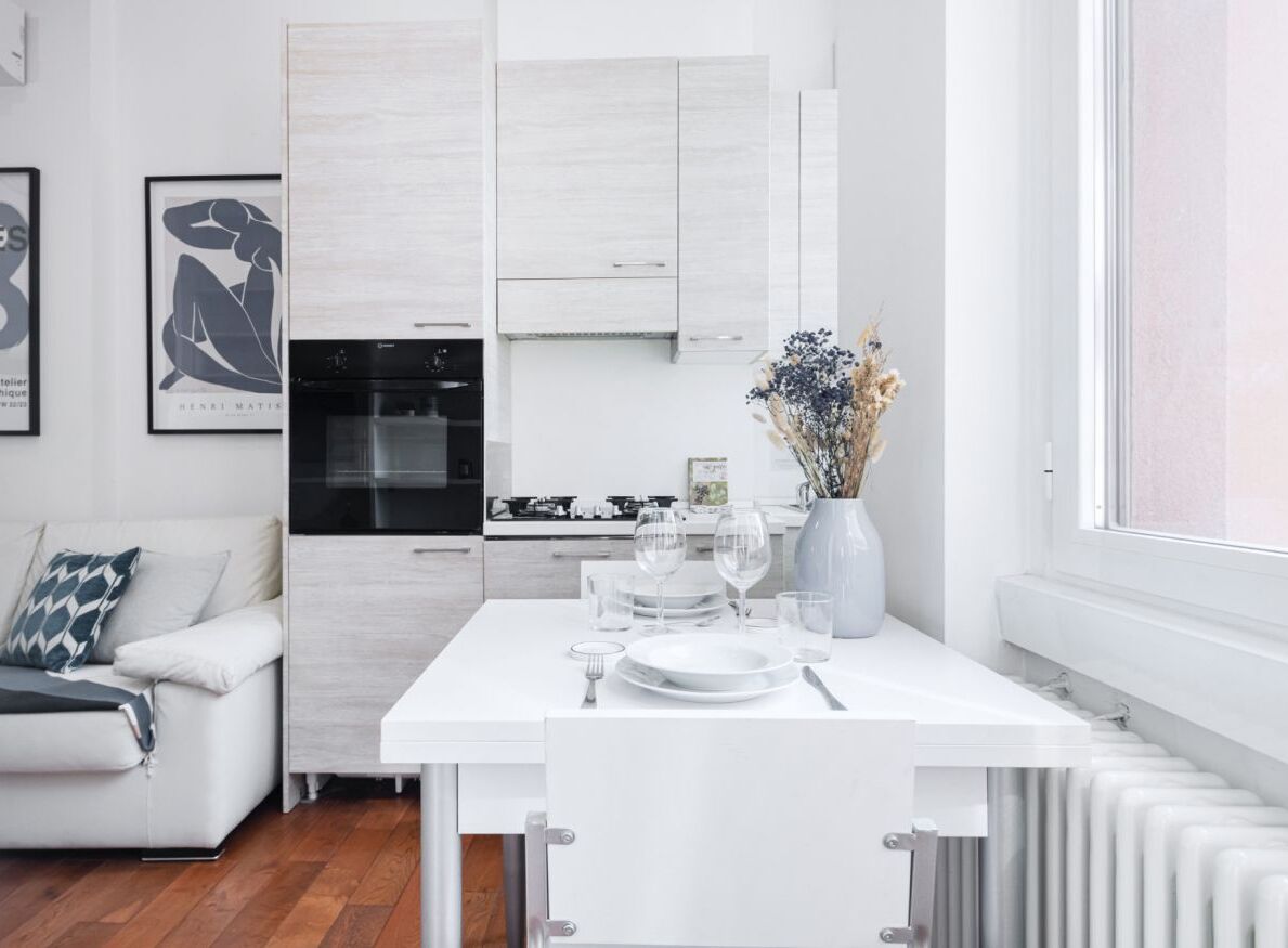 Een moderne keuken met witte kastjes, een eettafel gedekt met borden en glazen, en een vaas met gedroogde bloemen.