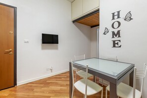 A room with a glass-top table, white chairs, a wooden door, a mounted TV, and a wall with 'HOME' and butterfly decals.