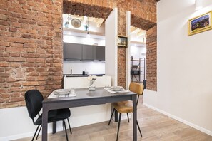A dining area with a brick wall, a modern kitchen in the background, and a wooden floor.