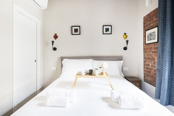 A neatly made bed with white linens, a tray with cups and a flower, and a brick wall with a picture.
