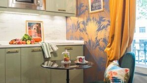 A compact kitchen with a microwave, a small table with a fruit bowl and a teapot, and a framed picture on the wall.