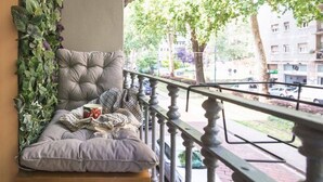 A balcony with a cushioned seating area, a railing, and a view of a tree-lined street.