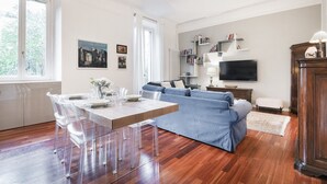 A modern living room with a wooden floor, a blue sofa, a dining table with clear chairs, and a TV mounted on the wall. - Easylife - Oasis of comfort&style in Porta Romana (Milano)