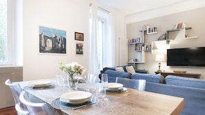 A modern living room with a wooden dining table set for four, a blue sofa, a flat-screen TV, and bookshelves. - Easylife - Oasis of comfort&style in Porta Romana (Milano)
