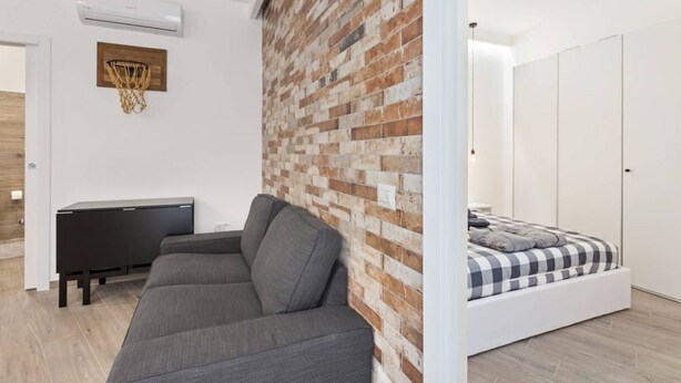 A modern bedroom with a brick accent wall, a bed with a checkered duvet, a grey sofa, a small bedside table, and a wall-mounted basketball hoop. - Easylife - Modern and new flat in Duomo area (Milano)