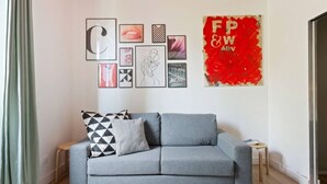 A grey sofa with patterned cushions, a small wooden stool, and a wall with various framed artworks and a red abstract piece.