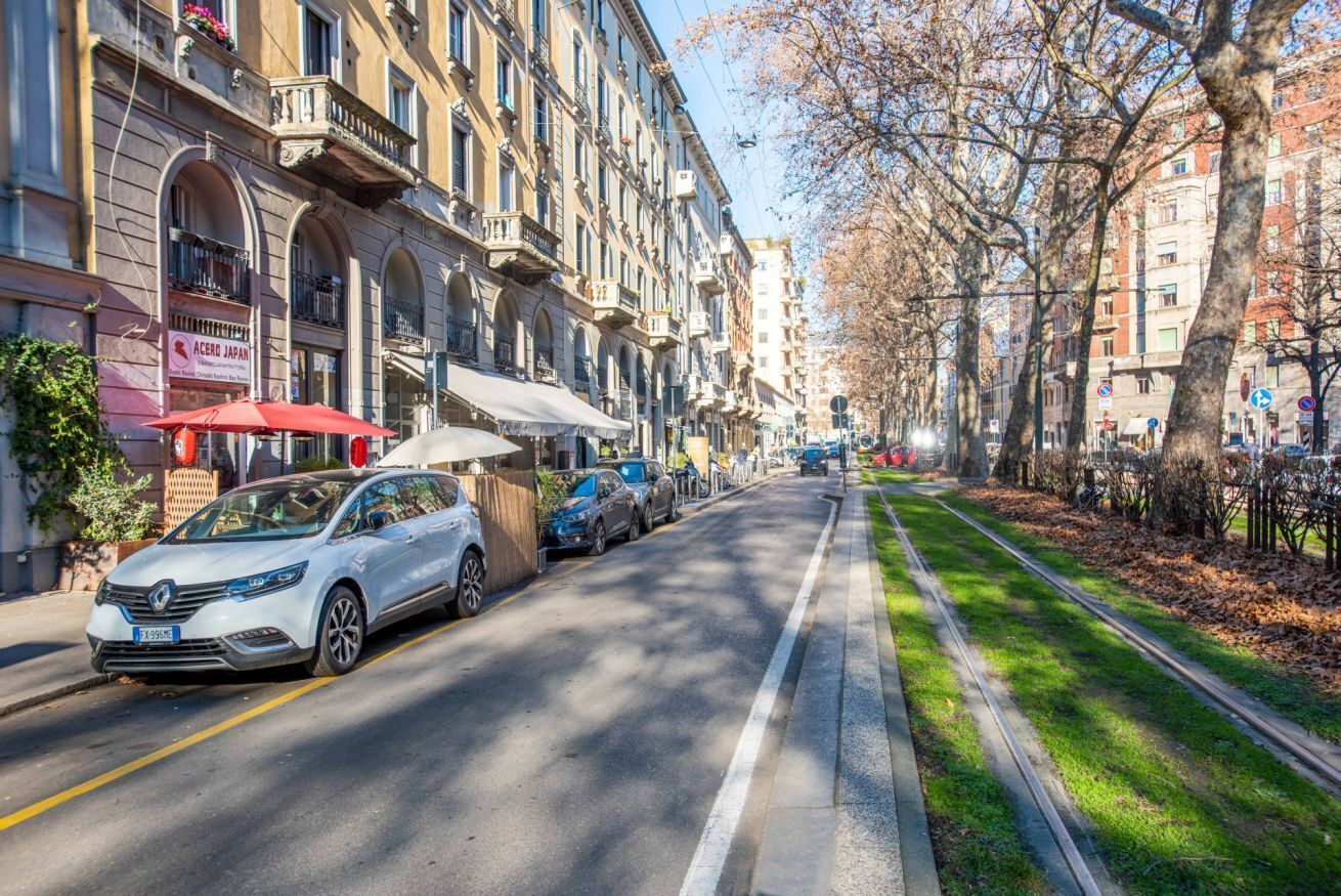 A street lined with multi-story buildings, parked cars, and a sidewalk with trees.