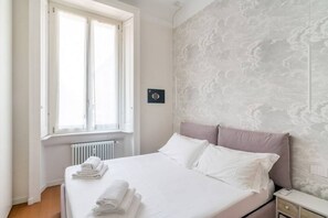 A neatly made bed with white linens and a folded towel, a bedside table with a lamp, and a window with curtains. - Easylife - Stylish and fancy flat in Buenos Aires (Milano)