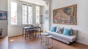 Amplia sala de estar con un sofá blanco, una mesa de comedor con sillas y un gran ventanal con vistas a edificios.