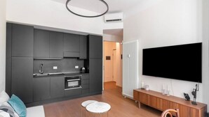 Una cocina moderna con armarios oscuros, un horno empotrado y un televisor de pantalla plana grande montado en la pared. Hay una zona de comedor con una mesa y sillas de madera, y un taburete pequeño y redondo.
