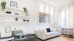 A modern living room with a white sofa, a dining table with chairs, a coffee table, and a bookshelf with decorative items.