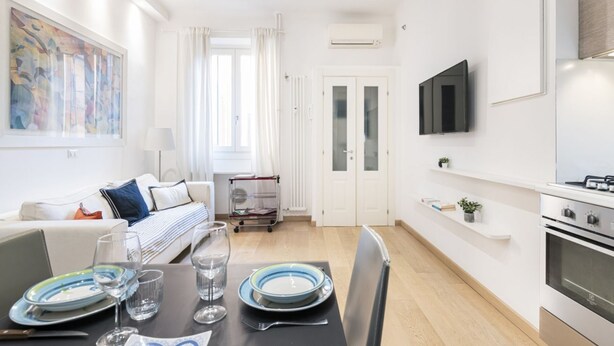 A modern, well-lit living room with a dining area, a sofa, a TV mounted on the wall, and a kitchen area with a stove.
