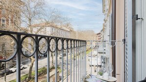 A balcony with a wrought iron railing, a table with a magazine, and a view of a street with buildings and trees.