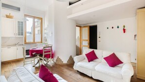 En moderne stue med en hvid sofa, et spisebord med stole, og et køkkenområde med hvide skabe og et vindue.