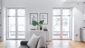 Un salon moderne avec un canapé gris, une petite table blanche ornée de deux tableaux encadrés et une grande fenêtre avec des stores.