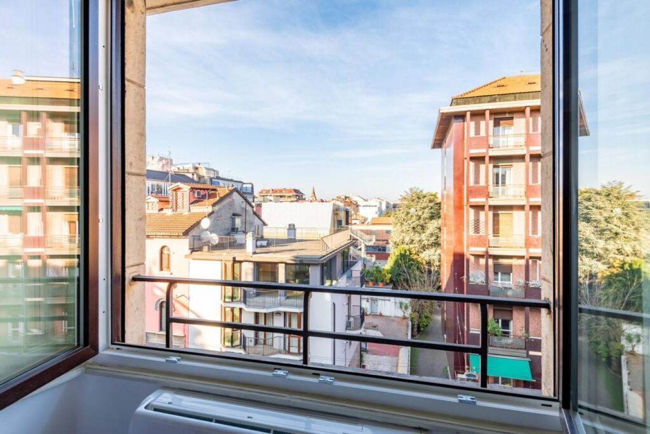Blick aus einem Fenster eines Wohnhauses mit mehreren Balkonen, einer sichtbaren Klimaanlage und blauem Himmel.