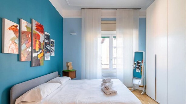 A bedroom with a bed, a nightstand, a window with curtains, and a wall with abstract paintings.