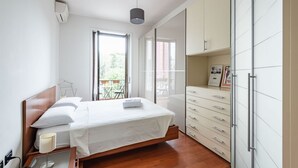 Un dormitorio moderno con cama de madera, ropa de cama blanca, lámpara de noche, armario con varios cajones y balcón con mobiliario exterior.