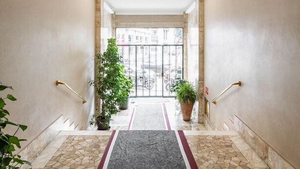 Entrada de hotel con piso de baldosas, un tapete central, plantas en macetas y una puerta de vidrio que da al exterior.