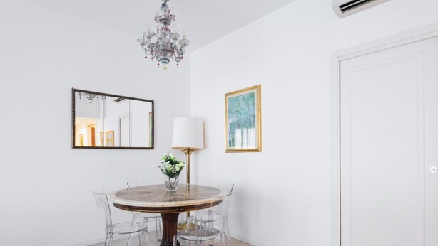 A dining area with a round table, clear chairs, a chandelier, a mirror, and framed artwork on the wall.