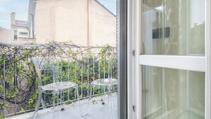 A balcony with a glass door, clear glass chairs, and a view of buildings and greenery.
