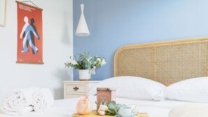 Ein ordentlich bezogenes Bett mit weißen Leinen, ein Korbschlafbrett, ein Nachttisch mit einer weißen Lampe, eine dekorative Vase mit Blumen, eine rosa Vase und ein Tablett mit Teekanne und Tasse.