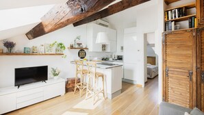 Eine moderne Küche mit weißen Schränken, einer zentralen Kochinsel und Holzbalken. Es gibt einen Essbereich mit Fernseher und einer Couch.