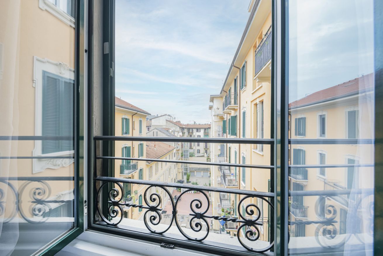 Una ventana con vistas a una calle estrecha flanqueada por edificios, un balcón con barandilla de hierro decorativa y un cielo despejado.