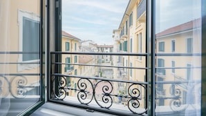 Une fenêtre donnant sur une rue étroite bordée d’immeubles, un balcon orné de balustrades en fer forgé et un ciel dégagé.