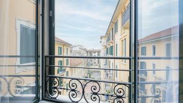 Una ventana con vistas a una calle estrecha flanqueada por edificios, un balcón con barandilla de hierro decorativa y un cielo despejado.
