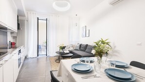 A modern kitchen with white cabinets and a red appliance, a dining area with a table set for two, and a living area with a sofa and a plant.