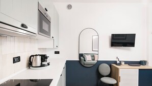 Une cuisine compacte équipée d’une cuisinière, d’un four à micro-ondes et d’une bouilloire, un petit coin salon avec un canapé et un fauteuil, et un téléviseur fixé au mur.