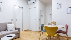 A modern living room with a sofa, a coffee table, and a dining table with chairs. There is a door leading to another room, an air conditioning unit on the wall, and a phone on the wall.
