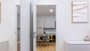 A modern hotel room with a wooden floor, a bed, a desk, and a chair. There is a kitchen area visible through an open door.
