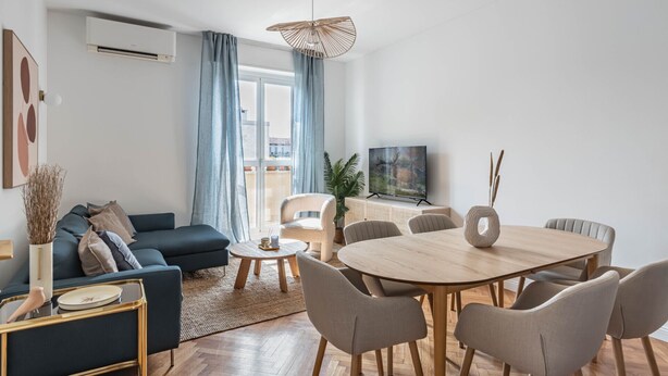 Un salon moderne avec un canapé bleu foncé, une table de salle à manger ronde en bois entourée de six chaises, un téléviseur à écran plat posé sur un meuble et une grande fenêtre ornée de rideaux bleus.
