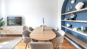 Un espace repas moderne avec une table en bois et des chaises à revêtement gris, un téléviseur posé sur un meuble blanc, et un mur d’accent bleu agrémenté d’étagères.