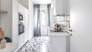 Une cuisine étroite avec des meubles blancs, un four encastré et un pare-feu en carreaux.
