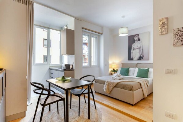 A modern apartment with a dining area, kitchenette, and a bedroom with a bed and pillows.