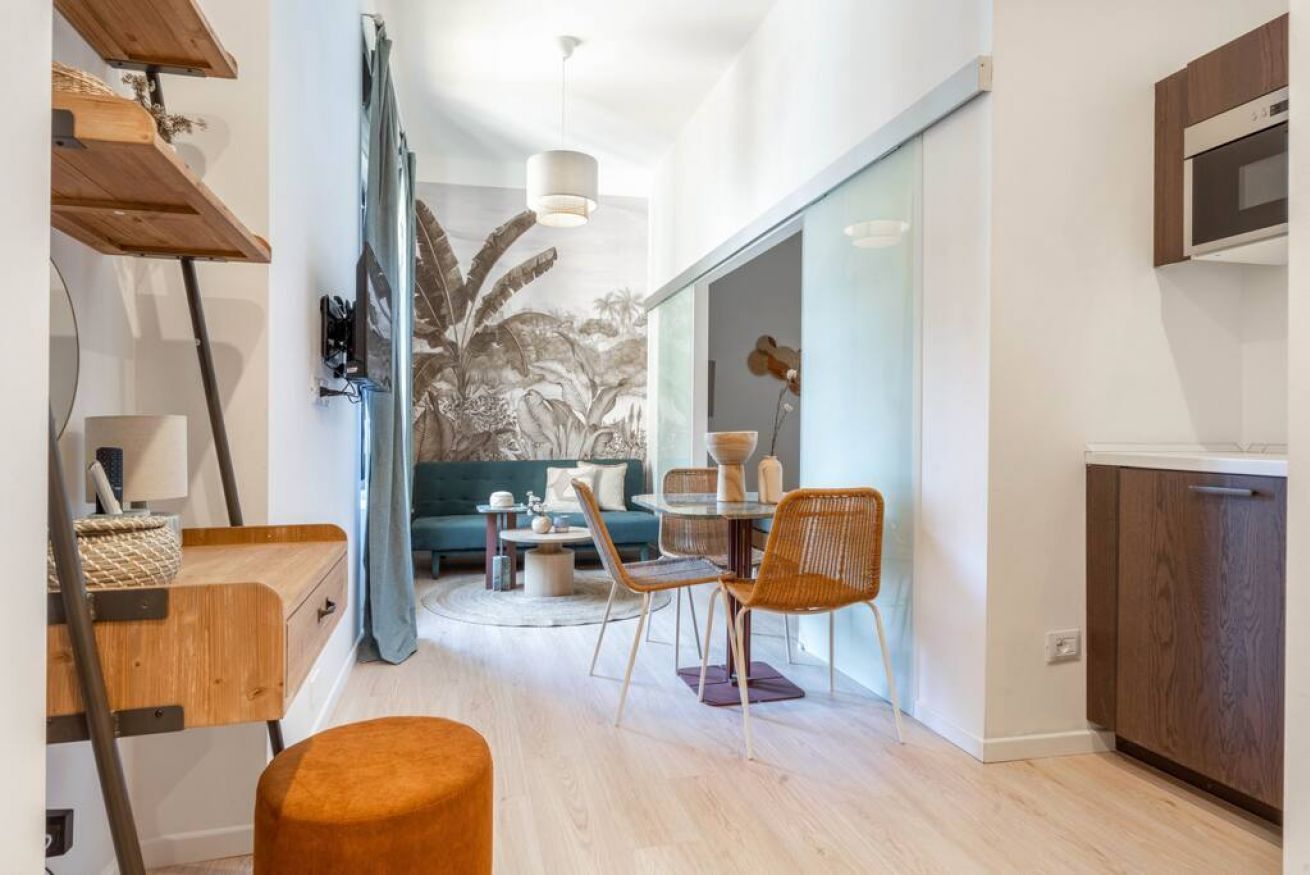 Eine moderne Küche mit Holzausstattung, ein Essbereich mit rundem Tisch und Stühlen sowie ein Wohnzimmer mit Sofa und einer großen Pflanze.