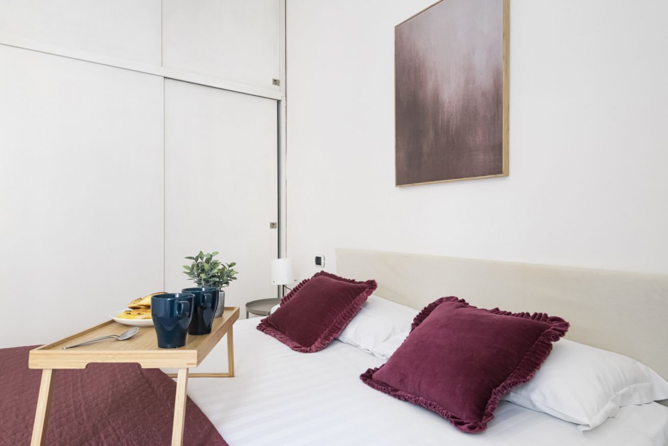 Een slaapkamer met een bed, twee kussens, een nachtkastje met dienblad en een aan de muur hangend schilderij.