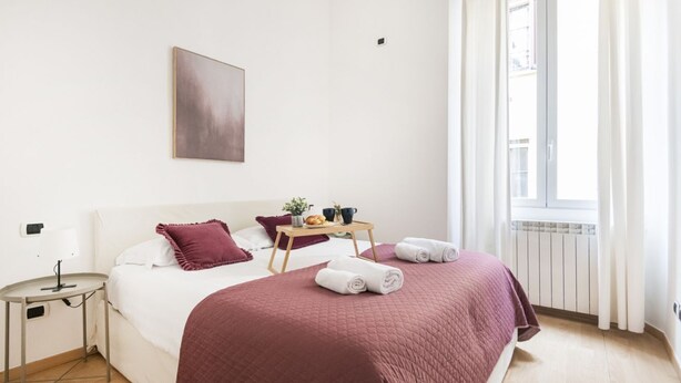 A neatly arranged bedroom with a bed, a bedside table, a small table with a tray, and a window with curtains. - Easylife - Comfortable three-room apartment in the heart of Milan (Milano)