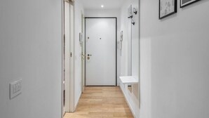 A narrow hallway with a wooden floor, white walls, and a white door at the end. There are framed pictures on the wall and a small white shelf on the right. - Easylife - Sophisticated studio with balcony in the heart of Porta Venezia (Milano)